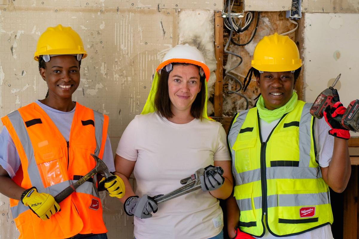 Three tradeswomen in hard hats and safety vests holding their tools